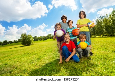 Group of very happy six boys, team sitting on the grass with balls and smiling, on sunny summer day