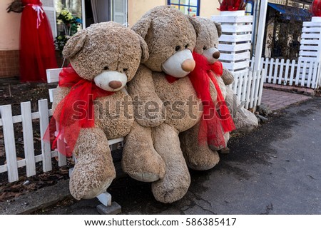 Group vintage soft toy teddy bear with red bows sit on bench in street near entrance to toy store. Old retro teddy bear sitting on bench in street. Soft toys as concept, symbol of love, care childhood