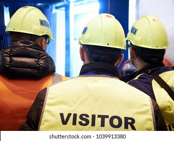 Group Of Visitors On The Factory Tour. People Go In Helmets And Uniforms For An Industrial Enterprise