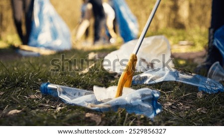 Group of volunteers cleaning the forest from litter and plastic waste, grabbing trash and junk with a long claw tool. Environmental activists picking up rubbish. Close up. Camera B.