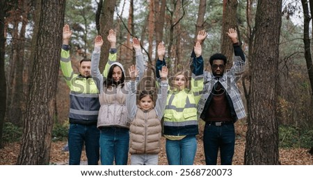 Group of volunteers looking at camera while standing in middle of forest. Raising hands above head level. Wearing yellow reflective vests. Bags with trash laying on ground in background.