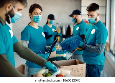 Group Of Volunteers Wearing Protective Face Masks While Packing Food And Clothes In Donation Boxes.