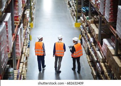 Group of warehouse workers wearing hardhats and reflective jackets waking in aisle between tall racks with packed goods, back view - Powered by Shutterstock