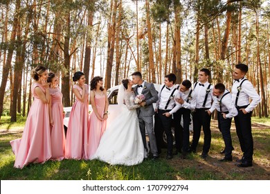 Group wedding photo. Bride and groom kisses near friends, bridesmaids in pink dresses and groomsmen with bow ties and suspender looks at newlyweds