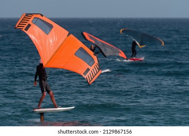 A Group Is Wing Foiling Using Handheld Inflatable Wings And Hydrofoil Surfboards In A Blue Ocean, Rider On A Wind Wing Board, Surf The Waves