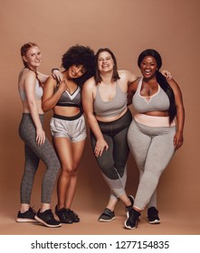 Group of women of different race, figure type and size in sportswear standing together over brown background. Diverse women in sports clothing looking at camera and laughing.