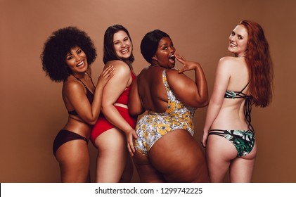 Group of women of different race, figure and size in swimsuits standing together and laughing against grey background. Diverse women in bikinis looking at camera.