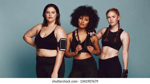 Group of women of different race, figure type and size dressed in sportswear standing together against grey background. Three diverse women in sports clothing looking at camera.
