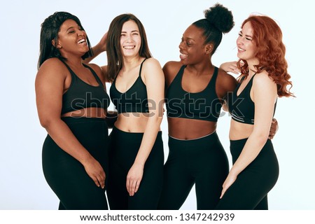 Group of women of different race, figure and size in sportswear standing together, chatting and laughing against white background.