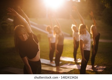Group of women do yoga in city park on summer sunny sunrise or sunset. Group of people are tilting to the side with their hands up