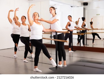 Group of women engaged in ballet in a dance studio perfoms an exercise near the ballet barre, where the choreographer helps ..to coordinate the movements correctly