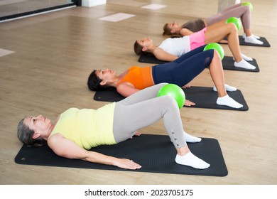 Group of women performing set of exercises with small pilates ball during group class in fitness studio
