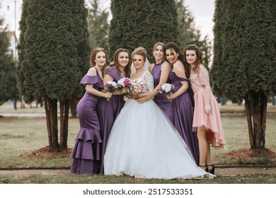 A group of women are posing for a picture in front of trees. One of the women is wearing a white dress and holding a bouquet of flowers. The scene has a festive - Powered by Shutterstock