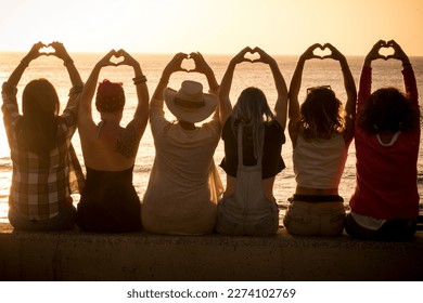 Group of women sitting against a golden sunset and doing heart symbol with hands and raised arms for friendship and love freedom concept. Women international day. Travel and vacation. Friends at beach