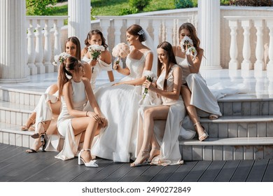 A group of women are sitting on steps, some of them are wearing wedding dresses. They are holding flowers and posing for a picture. Scene is joyful and celebratory - Powered by Shutterstock