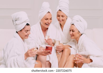 A group of women spend leisure time. Four Young attractive women in bathrobes look at the smartphone screen and smile. - Powered by Shutterstock