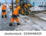 A group of workers are clearing snow from a street. The workers are wearing orange jackets and are using shovels and a large snow plow. The scene is cold and snowy