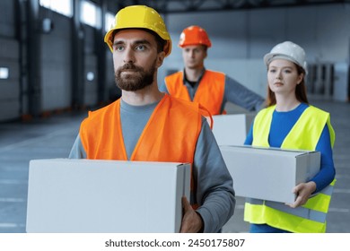 Group of workers, loaders, wearing hard hats and work uniforms, hold boxes, mockup looking away. Attractive bearded man carries box to warehouse. Concept of logistics, delivery - Powered by Shutterstock