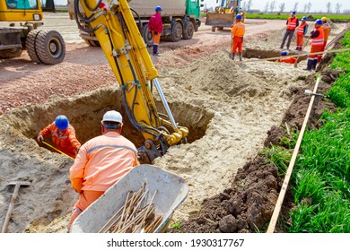 Group Of Workers With Safety Vests And Helmets Are Help Excavator To Excavating Square Trench In Soil At Construction Site.