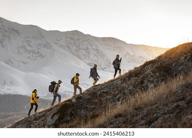 Group of young active hikers with backpacks walks uphill in mountains at sunset