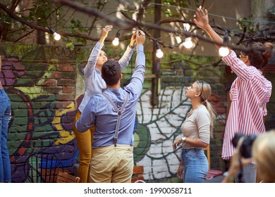 group of young adult caucasian friends setting up hanging bulbs on branches in the garden, preparing for party