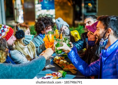group of young adults celebrating christmas holidays toasting together in a party at night after coronavirus lockdown reopening.happy friends wearing protective face mask in open bar restaurant.