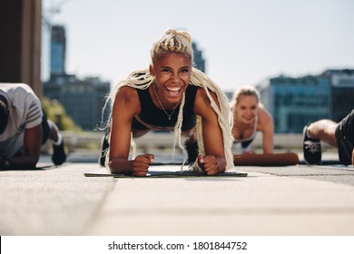 Group Of Young Adults Working Out Together Outside In The City. Men And Women Holding A Plank Position And Smiling.