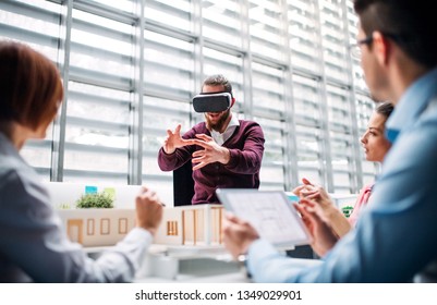 Group Of Young Architects With Model Of A House And VR Goggles Working In Office.