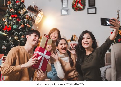 Group young Asian friends celebrating Christmas and New Year in the evening, sitting holding gift boxes taking a selfie together on the smart phone near the Christmas tree at home. - Powered by Shutterstock