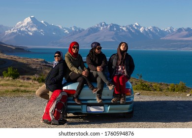 A Group Of Young Asian Traveller Friends On A Roadtrip Through Countryside, Sitting On Rear Car Boot, Smile And Happy Face. Mountains In The Background
