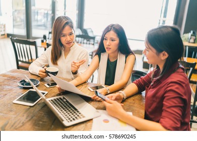 Group Of Young Asian Women, College Students In Serious Business Meeting Or Project Brainstorm Discussion At Coffee Shop. With Laptop Computer, Digital Tablet, Smartphone. Startup Or Teamwork Concept.