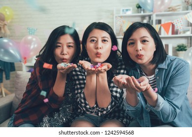 Group Of Young Asian Women Holding Colorful Confetti On Hands Blowing Sitting On Couch. Best Friends Having Fun In Decorated Living Room At House Party. Three Charming Girls Having Fun Indoors.