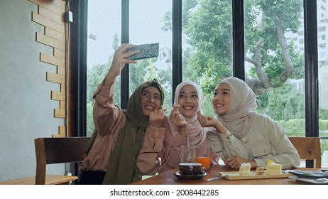 Group of young beautiful muslim women wearing hijab sitting near glass window in coffee shop, they are use smartphone to selfie with happiness - Powered by Shutterstock