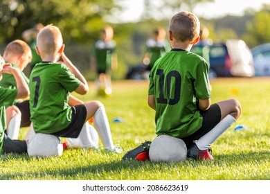 Group Of Young Boys Kicking Sports Soccer Game On Grass Pitch. Boys Sitting On Soccer Balls Waiting To Play The Game. Friends In Sports Team. School Boys On Sideline. Kids In Green-Sports Shirts