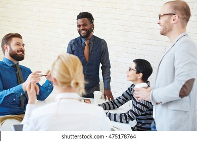 Group Of Young Business Men And Women Meeting In Office, Talking And Telling Stories And Jokes At Coffee Break, African American Man Standing Among Them Smiling Brightly