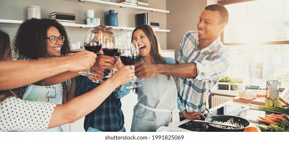 Group of young cheerful friends cheering with wine glasses while cooking some tasteful food at kitchen.