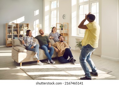 Group of young cheerful friends have fun playing charades during casual gathering at home. Man shows pantomime to people sitting in front of him on sofa. Friendship, leisure and entertainment concept.