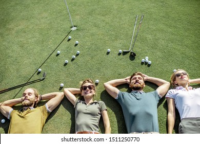 Group Of A Young And Cheerful Friends Lying On The Golf Course With Balls And Putters On The Grass, Resting And Having Fun After The Game