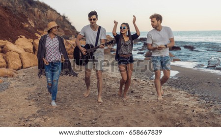 group of young and cheerful friends walking on beach, One man is playing guitar. Dancing and singing on wild beach. Happy friends