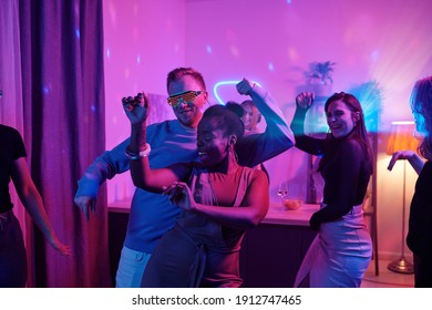 Grupo de jóvenes amigos interculturales alegres en ropa informal inteligente bailando excitados en fiestas en casa en el salón iluminado con iluminación rosa