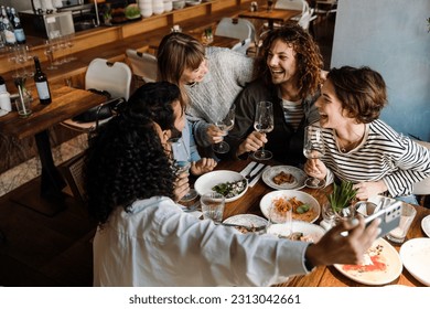 Group of young cheerful multinational friends taking selfie and drinking wine while dining at table in restaurant - Powered by Shutterstock