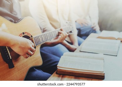 A group of young Christian friends plays guitar and sing a song to praise and worship God together with a bible and hymn song book on wooden table, Christian fellowship in small group concept - Shutterstock ID 2244793577