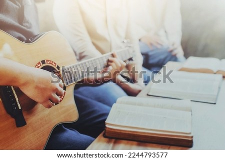 A group of young Christian friends plays guitar and sing a song to praise and worship God together with a bible and hymn song book on wooden table, Christian fellowship in small group concept