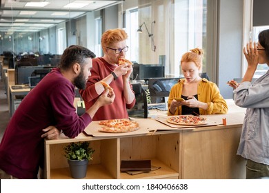 Group of young colleagues in casualwear talking and using smartphone at lunch break while having pizza by table in large contemoprary office - Shutterstock ID 1840406638