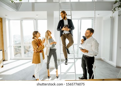 Group Of A Young Creative Office Employees Having Some Informal Discussion, While Standing Together In The Bright Meeting Room With Large Windows