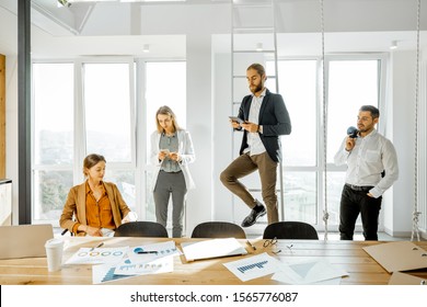 Group Of A Young Creative Office Employees Having Some Informal Discussion, While Standing Together In The Bright Meeting Room With Large Windows