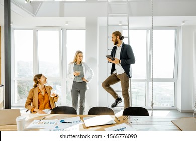 Group Of A Young Creative Office Employees Having Some Informal Discussion, While Standing Together In The Bright Meeting Room With Large Windows