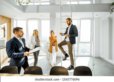 Group Of A Young Creative Office Employees Having Some Informal Discussion, While Standing Together In The Bright Meeting Room With Large Windows