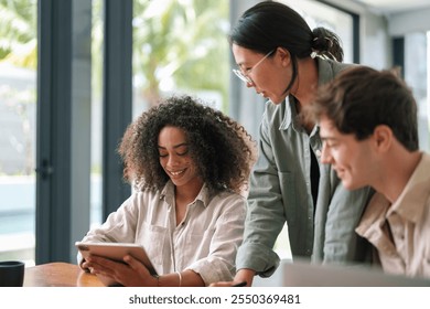 A group of young, diverse executives in casual business attire engage with a digital tablet as they collaborate on a project in a well-lit modern office environment. - Powered by Shutterstock