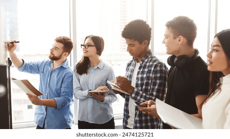 A group of young, diverse professionals are actively participating in a collaborative brainstorming session. One of them is writing on a whiteboard, while the others hold papers and digital devices - Powered by Shutterstock